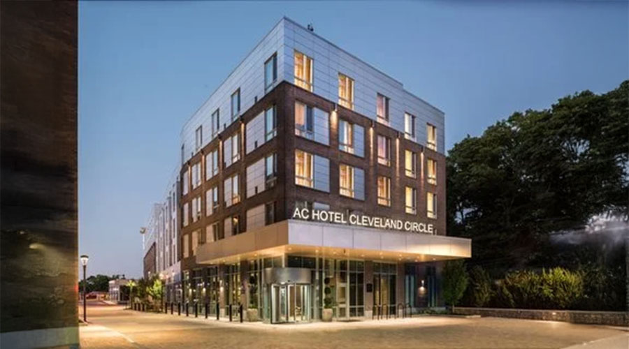 A modern four-story building with large windows and a glass entrance, labeled "AC Hotel Cleveland Circle," is illuminated at dusk, with trees and a quiet street in the background.