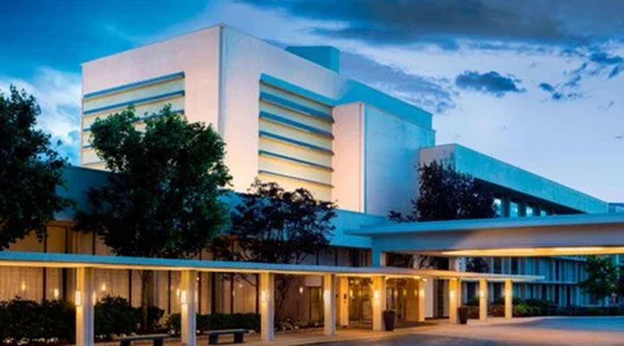 A modern building with white walls and blue accents is surrounded by trees and illuminated by outdoor lighting at dusk, with a covered walkway in the foreground.