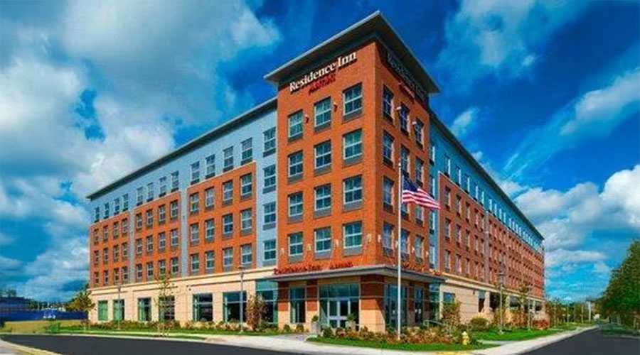 A modern, multi-story Residence Inn hotel with red brick and blue-gray exterior walls, large windows, and an American flag flying in front on a sunny day with a partly cloudy sky.