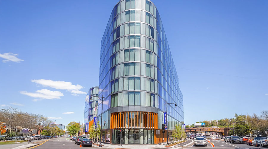 A modern, glass-fronted building with a curved facade stands at a street corner under a clear blue sky, surrounded by cars, trees, and other urban structures.