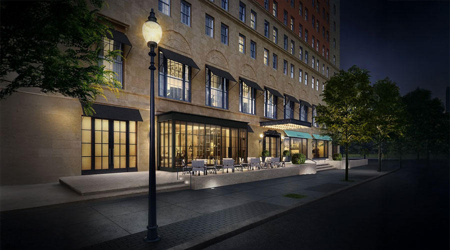 A well-lit hotel entrance at night features large windows, black awnings, an outdoor seating area, and a sidewalk lined with trees and a vintage streetlamp.